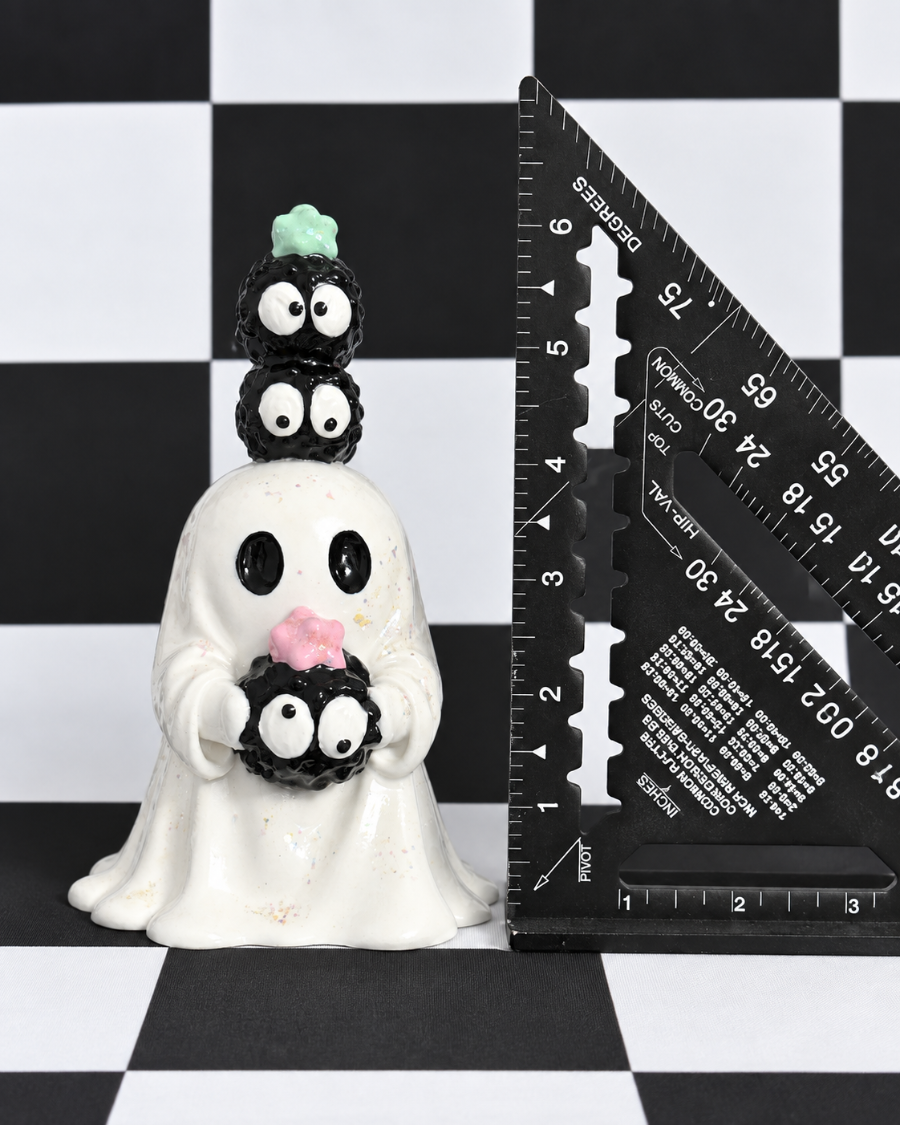 A small white ghost figurine with three stacked black-and-white spider characters on its head and a pink spider at the bottom, shown next to a black triangular ruler on a black-and-white checkered surface.