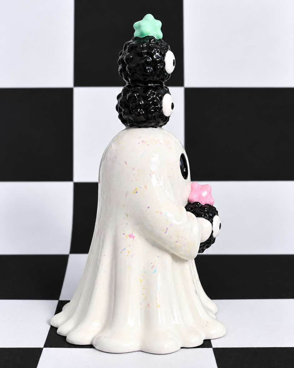 A ceramic ghost figurine with a black spiky head, white body, and holding a pink cupcake, set against a black and white checkered background.
