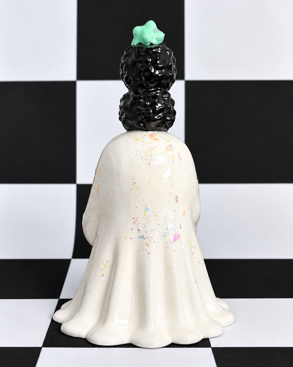 A ceramic ghost figurine shown from the back, featuring a cream-colored body with subtle pastel speckles, a black textured hair bun, and a mint green star on top, set against a black and white checkered background.