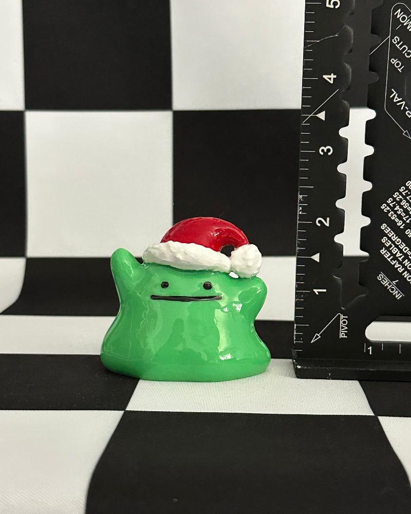 Green ghost figurine with a Santa hat on a black and white checkered background with a ruler for scale.