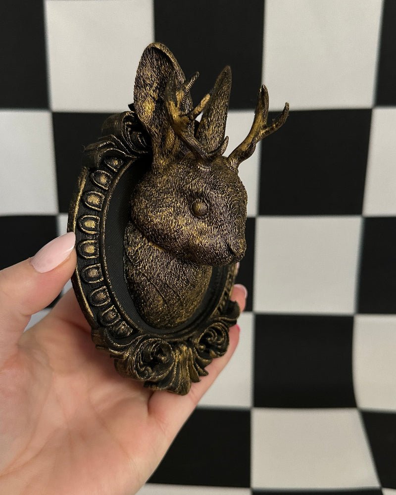 Decorative deer head with antlers in a bronze frame held against a black and white checkered background.