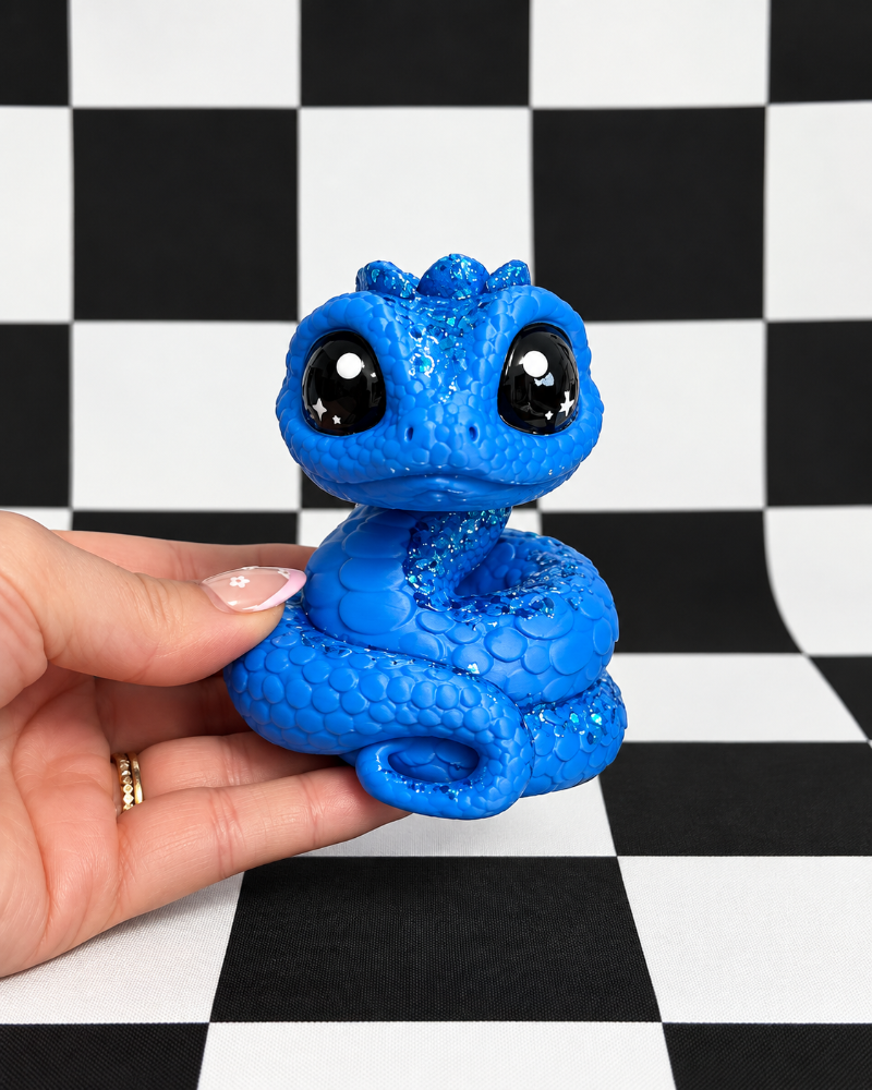 A small blue glittery snake figurine with large black eyes is being held in a person's hand against a black and white checkered background.