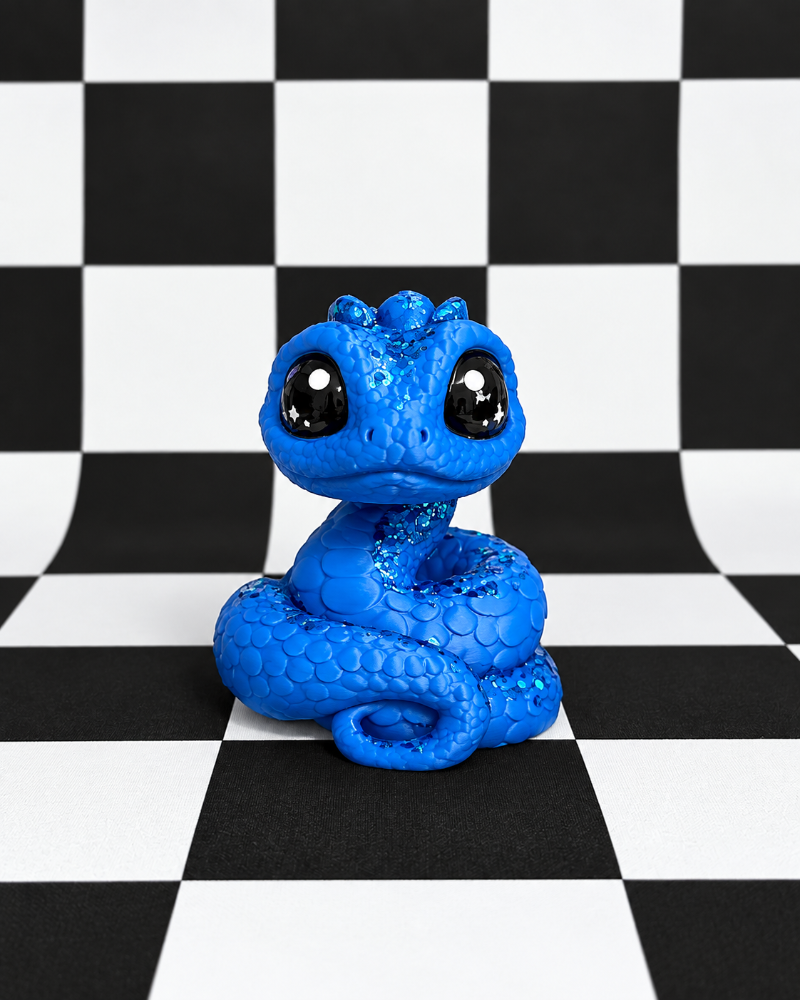 A bright blue coiled snake figurine with large glossy black eyes, centered on a black-and-white checkered surface and backdrop.
