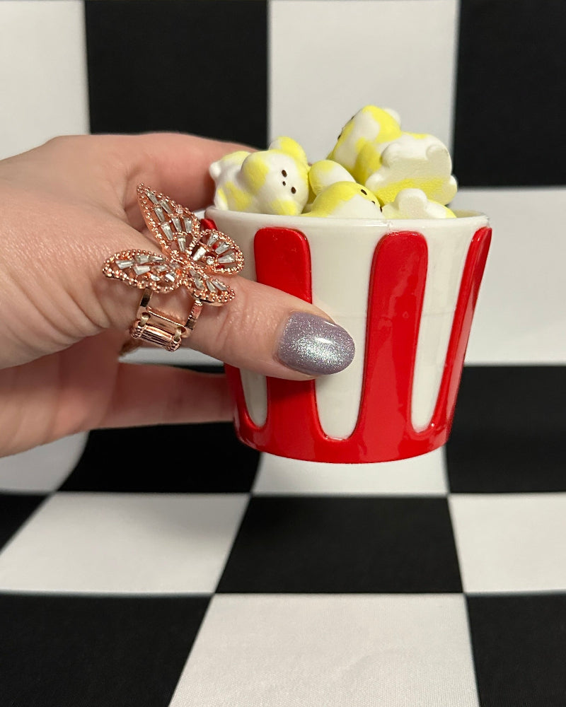 Hand holding a small red and white container filled with popcorn against a black and white checkered background.
