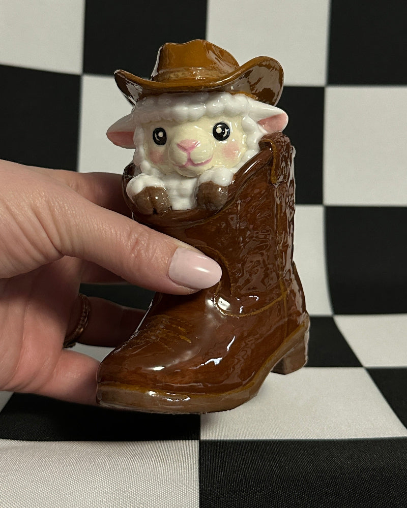 Small sheep figurine in a cowboy hat and boots held by a hand on a checkered background