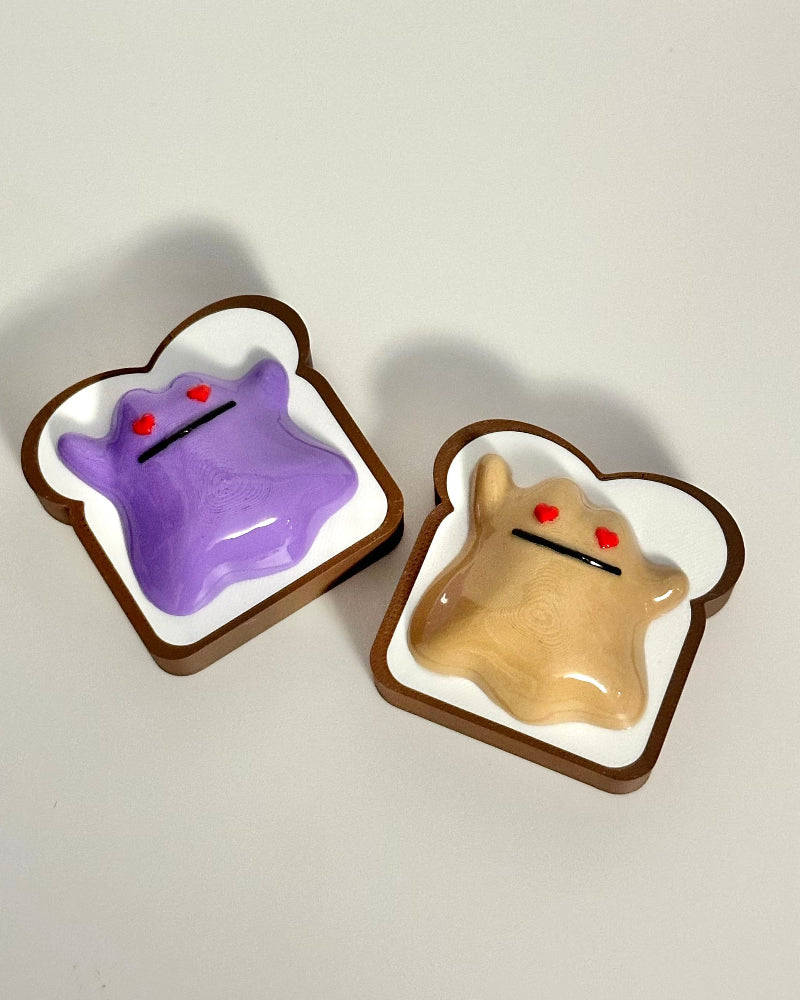 Two small figurines of a purple character and a beige character inside toast-shaped containers on a white background.