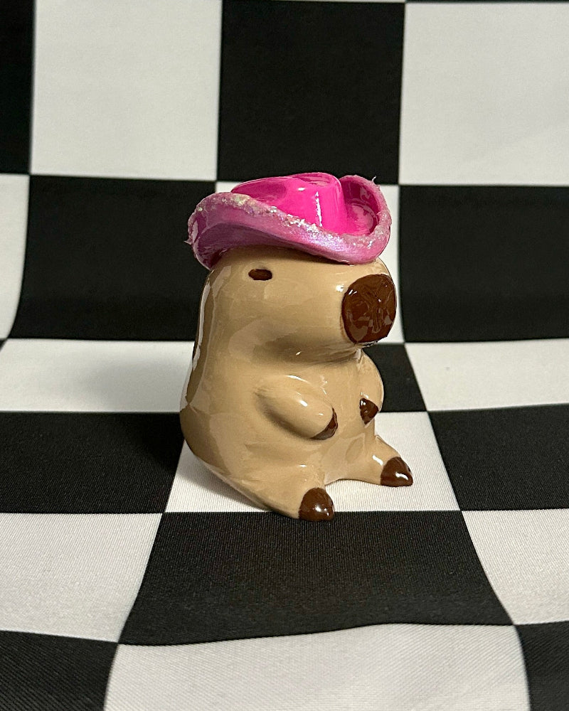 Small figurine of a capybara wearing a pink hat on a black and white checkered background