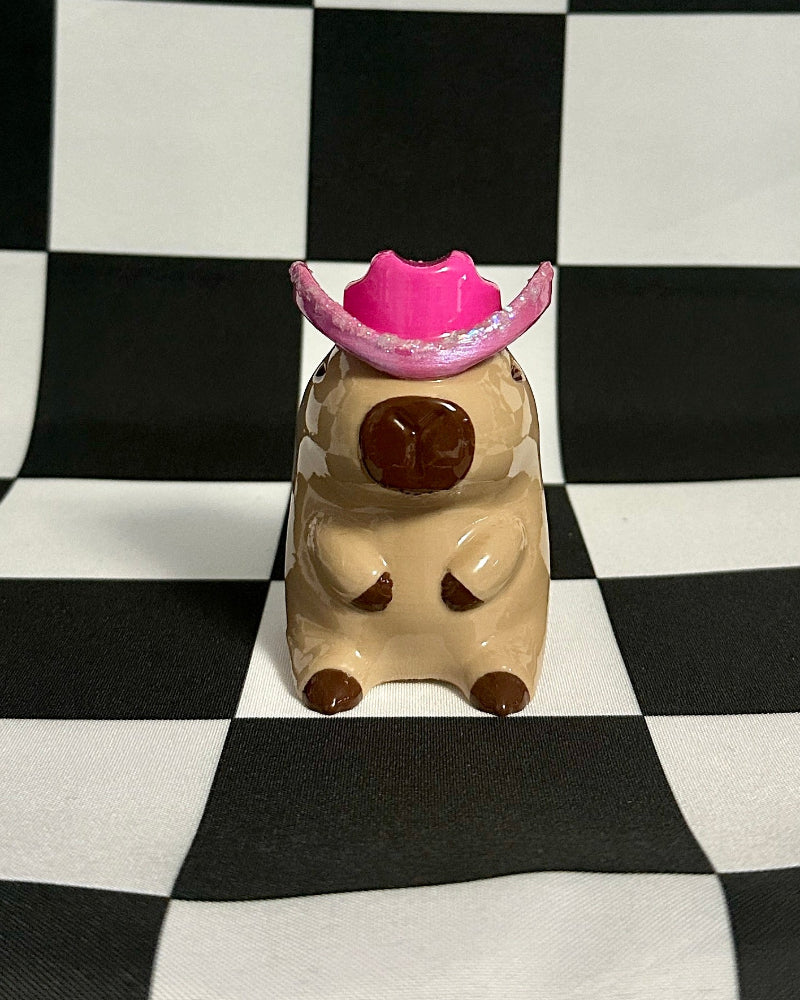 Small capybara figurine with a pink hat on a black and white checkered background