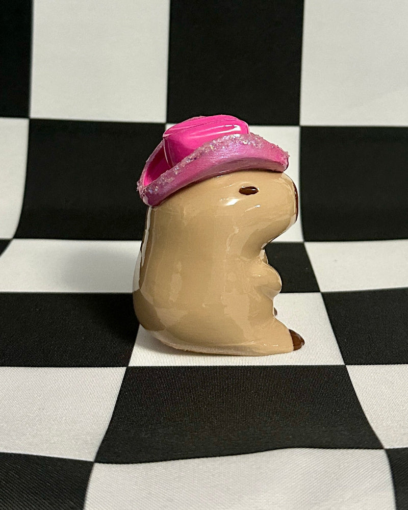 Small capybara figurine with a pink hat on a black and white checkered background