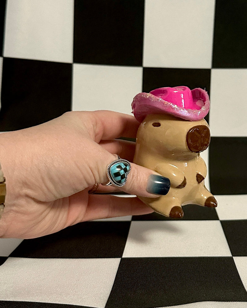 Small figurine of a capybara with a pink hat held by a hand against a black and white checkered background.