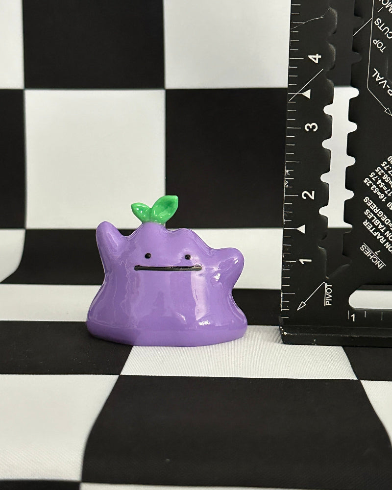 Purple cartoon-like creature with a green leaf on a black and white checkered background with a ruler for scale.