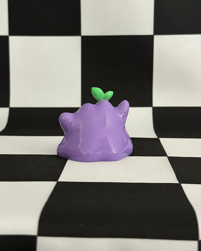 Purple creature figurine with a green leaf on a black and white checkered background