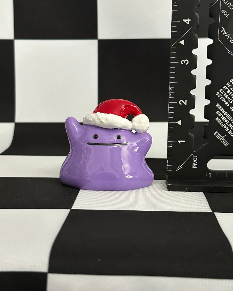 Purple character figurine with a Santa hat on a black and white checkered background with a ruler for scale.
