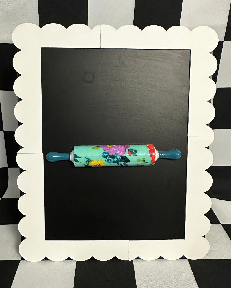 Floral-patterned rolling pin on a blackboard with a decorative white frame against a black and white checkered background.