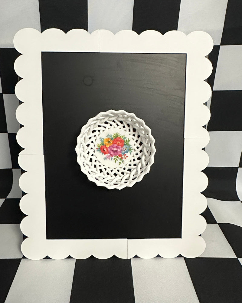 Decorative doily with floral design on a black background framed by a white scalloped frame against a black and white checkered background.