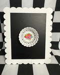 Decorative doily with floral design on a black background framed by a white scalloped frame against a black and white checkered background.