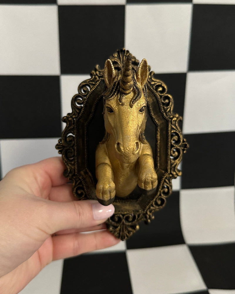 Gold unicorn figurine in a decorative frame held against a black and white checkered background