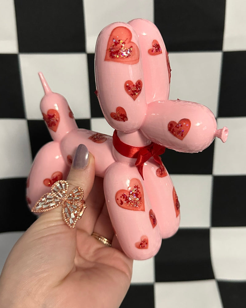 Pink balloon dog with heart patterns held by a hand against a checkered background