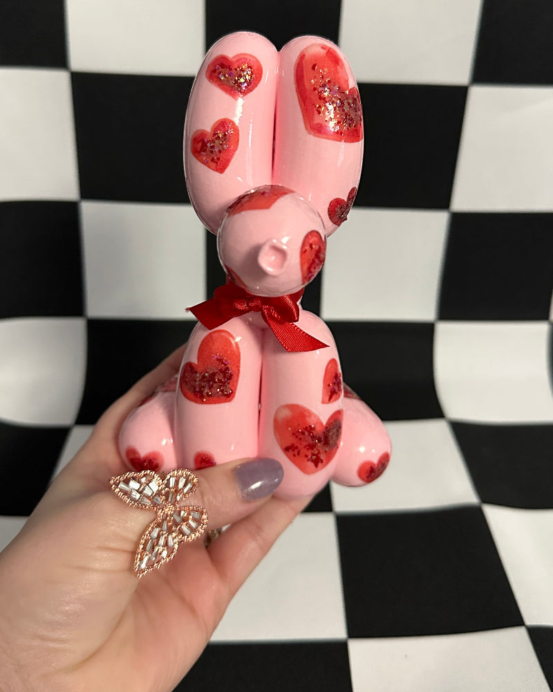Pink bunny figurine with red hearts and a bow tie held by a hand on a black and white checkered background.
