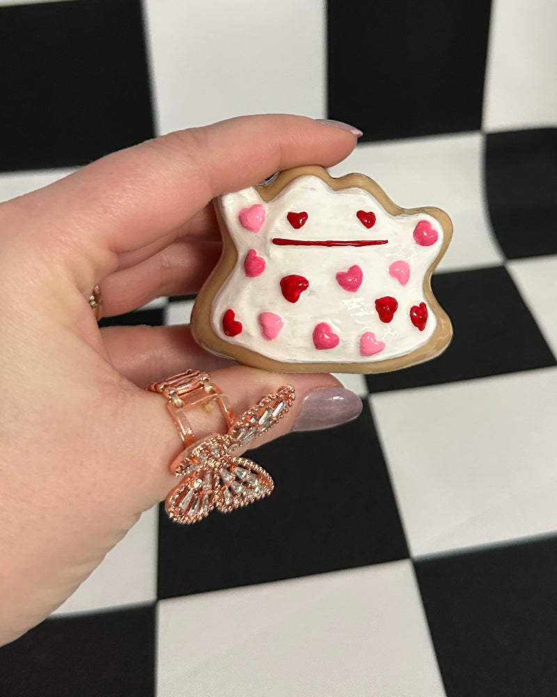 Hand holding a small cake-shaped cookie with red and pink decorations against a black and white checkered background.